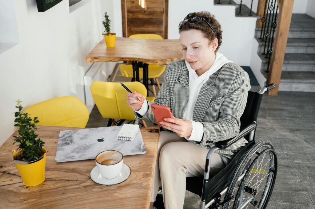 Cliente AccessiWeb Conseil fauteuil roulant consulte son smartphone à une table de travail, avec un ordinateur portable et un café à côté d'elle.